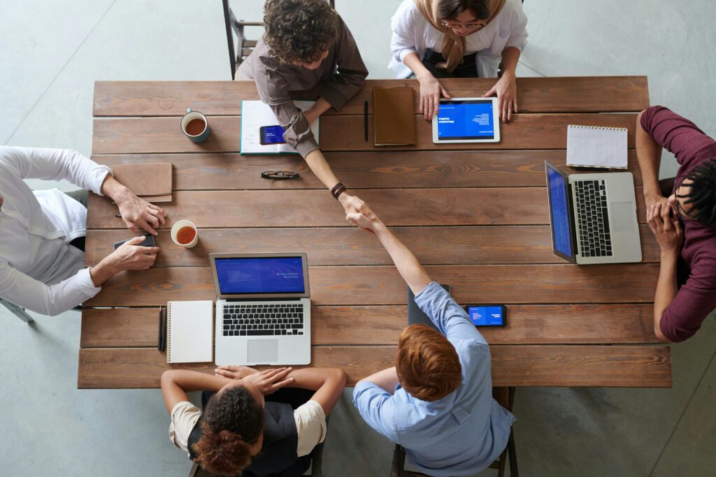 The purpose of this image is to visually represent the collaborative and team-oriented nature of modern tech workplaces, supporting your blog post about essential soft skills for tech professionals. It illustrates several key points from your article:

Teamwork and collaboration: The image shows a diverse group working together at a shared table.
Communication skills: Two people are shaking hands, symbolizing effective interpersonal communication.
Adaptability: The mix of traditional (notebooks) and modern (laptops, tablets) tools suggests adaptability to different work methods.
Problem-solving: The collaborative setup implies joint problem-solving and idea sharing.
Soft skills in tech
Tech professional development
Workplace collaboration
Communication in IT
Tech team dynamics
Problem-solving skills
Adaptability in technology
IT career growth
Tech industry teamwork
Professional skills for developers
Tech workplace culture
Interpersonal skills in IT
Tech talent management
Digital workforce skills
Tech industry best practices